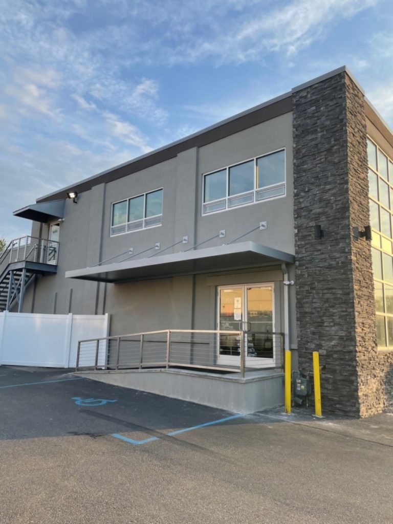 Modern building exterior with large windows, stone accents, and an accessible ramp at the entrance under a blue sky.