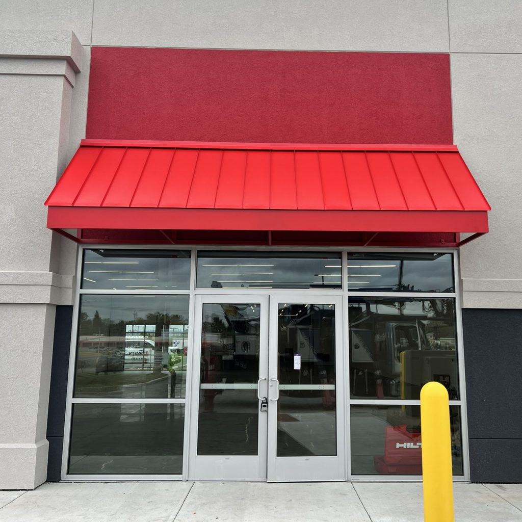 Red standing seam awning over glass entrance of a building with gray walls and a yellow post in front.