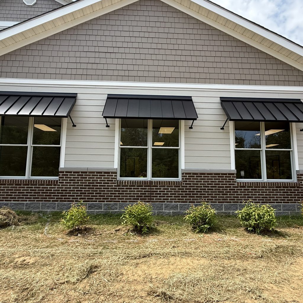 Exterior view of a building featuring several large windows, black awnings, and landscaped plants in front.